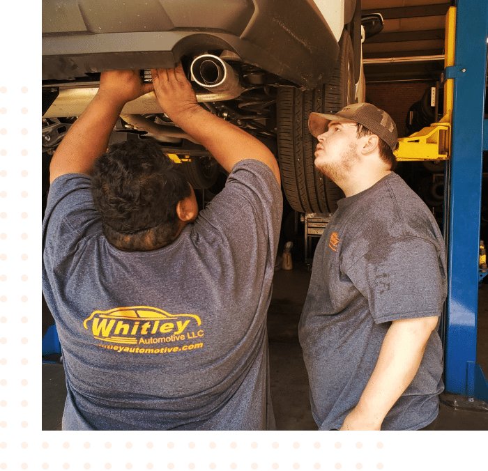 two male technicians work together on the exhaust of a vehicle that is raised on a lift in the shop