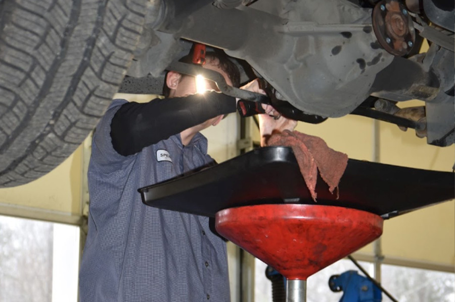 Guide on when to change your car’s fluids in Locust, NC. Image of Whitley Automotive technician standing under vehicle on lift in shop to change the transmission fluid and the drain they use to catch the fluid with.