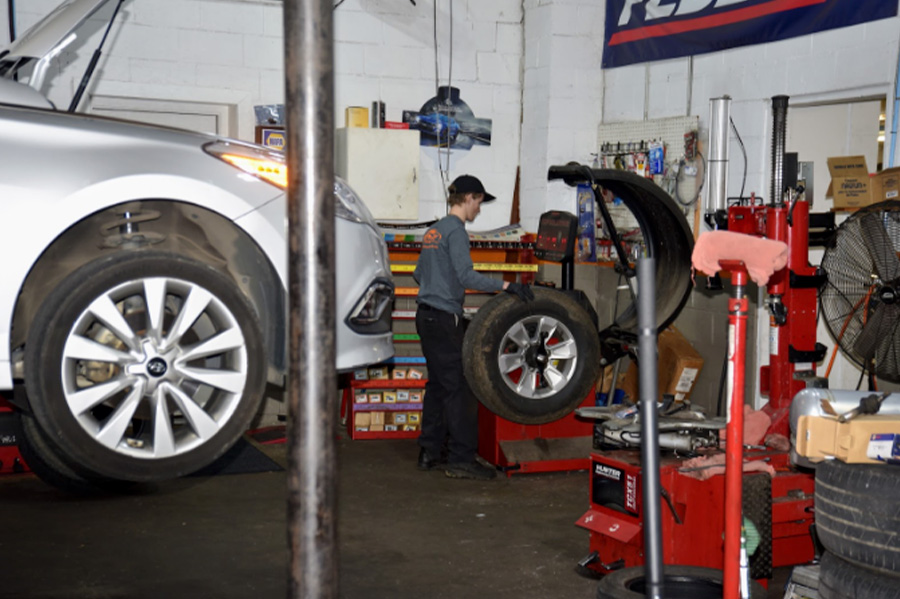 The importance of factory-recommended maintenance in Marshville, NC. Image of Whitley Automotive technician performing a tire rotation as routine maintenance for customer in shop near silver sedan on lift and shop tools