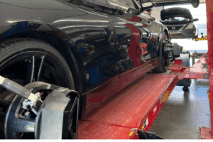 Wheel Alignment in Locust, NC by Whitley Automotive. A black car sits on an alignment rack inside an auto repair shop, with wheel alignment sensors attached to the front wheels. In the background, another vehicle is lifted with its hood open as a technician works.