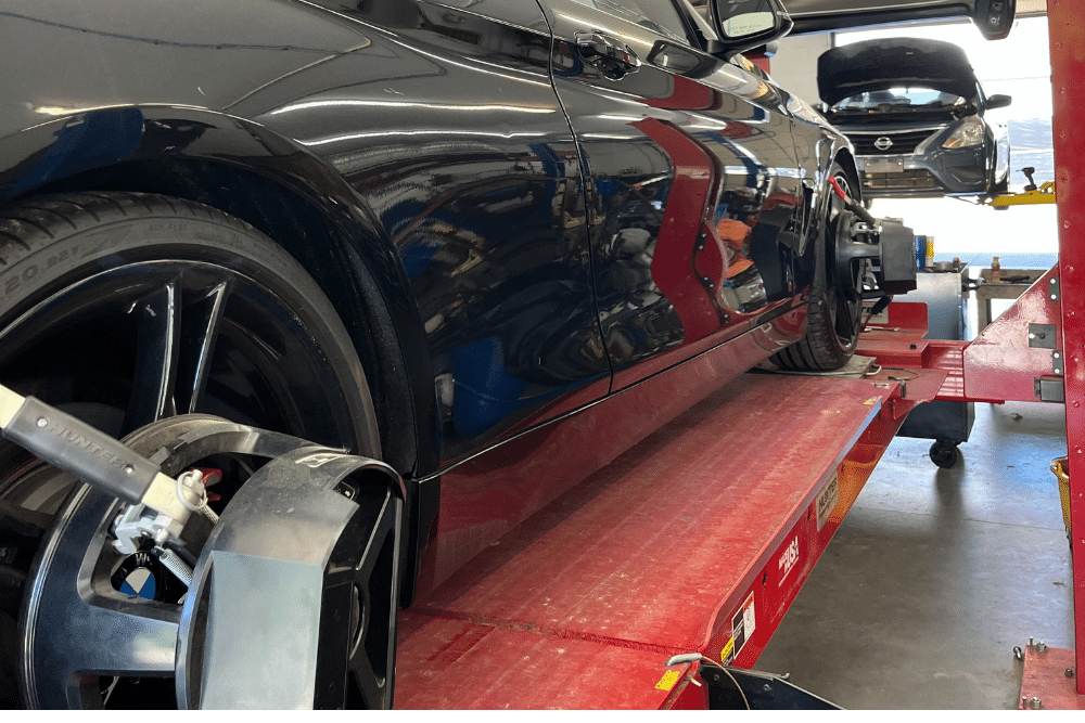 Wheel Alignment in Locust, NC by Whitley Automotive. A black car sits on an alignment rack inside an auto repair shop, with wheel alignment sensors attached to the front wheels. In the background, another vehicle is lifted with its hood open as a technician works.